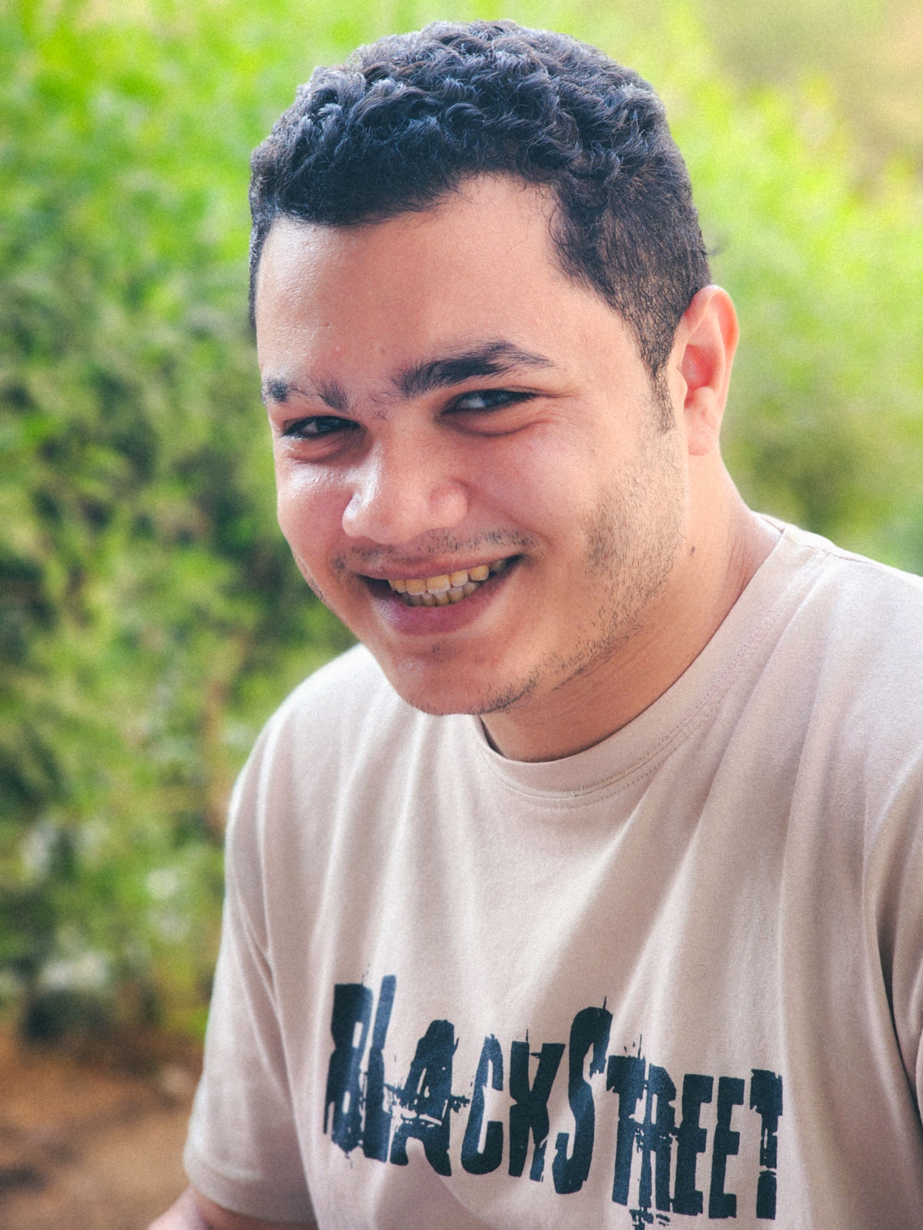 Cheerful young man with short curly hair smiling in a natural outdoor setting.