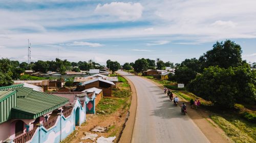 Free Aerial shot of motorcycles riding through a rural Cameroonian village road under a clear blue sky. Stock Photo
