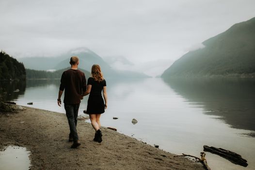 A couple walking hand in hand along a foggy lakeside path, mountains in the background.