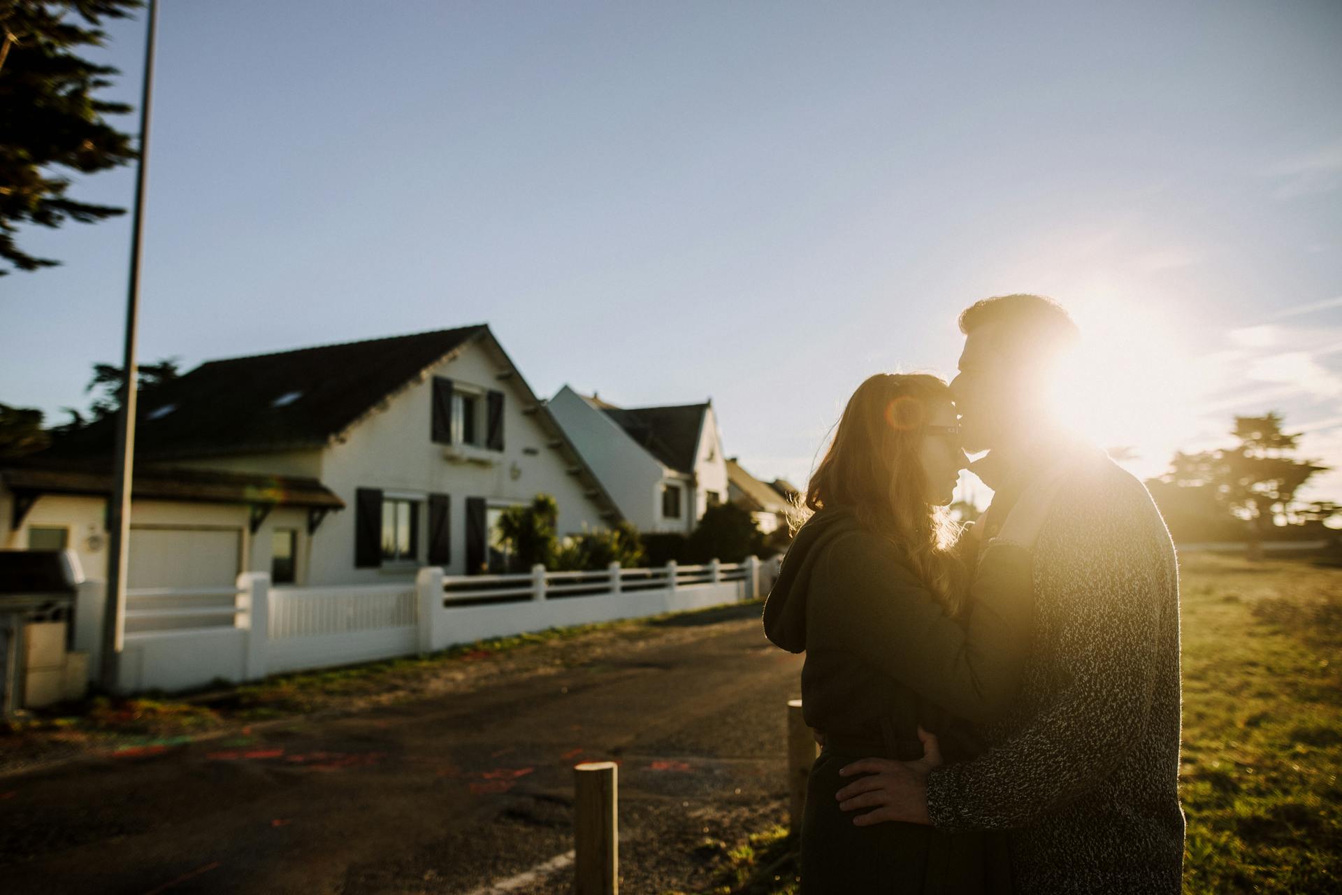 A couple shares a romantic embrace at sunrise in front of a cozy suburban house.