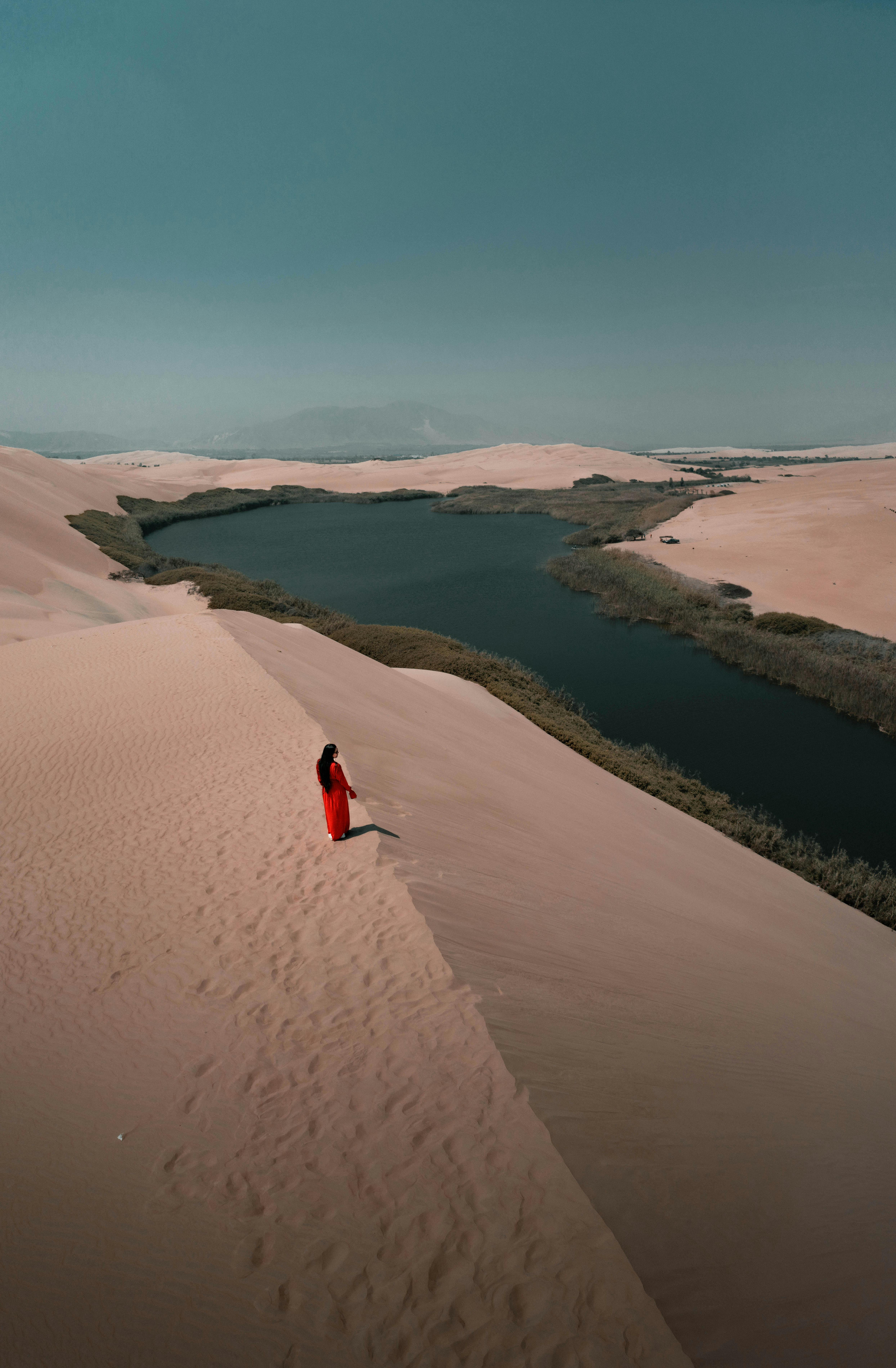Aerial view of a serene oasis in the Pisco desert with a woman in red, under a clear sky in Peru.
