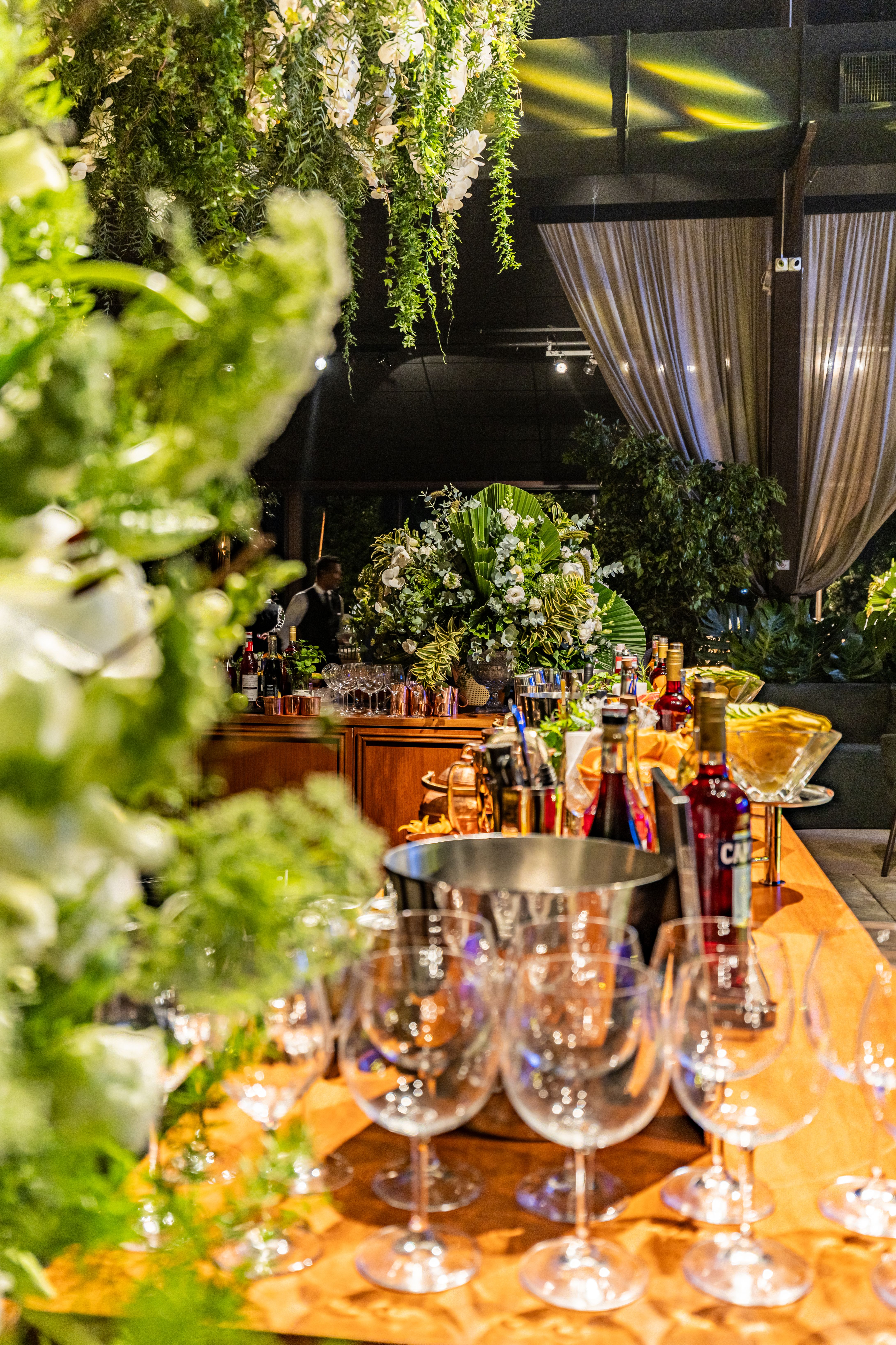 A sophisticated indoor event setting featuring lush greenery and elegantly arranged wine glasses on a wooden table.