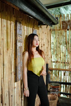 Stylish young woman in casual attire standing by a bamboo wall outdoors.