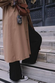 A stylish woman in a coat and sneakers ascending marble steps holding a vintage camera.