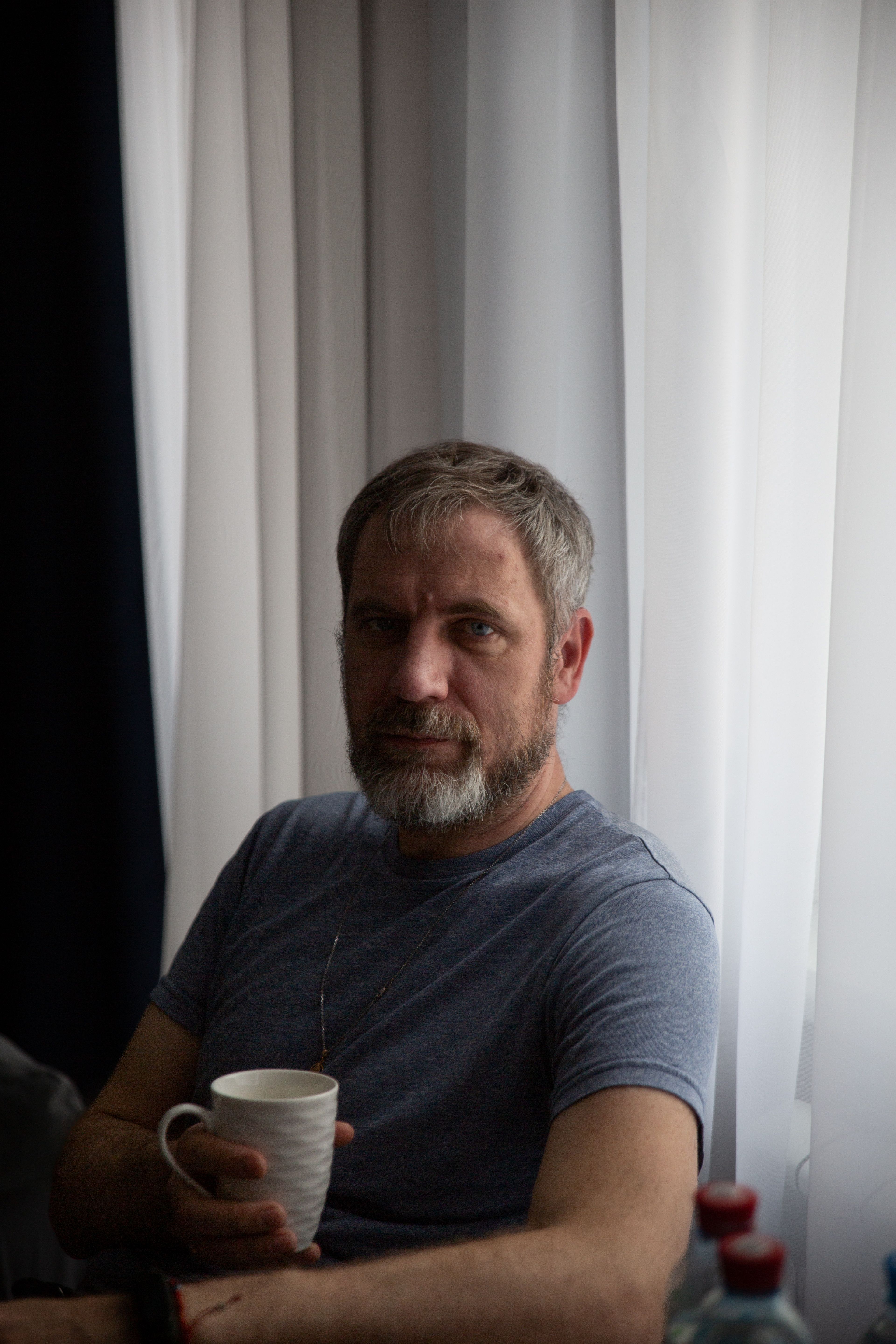Middle-aged man with a beard holding a mug, relaxing indoors by a window in soft light.