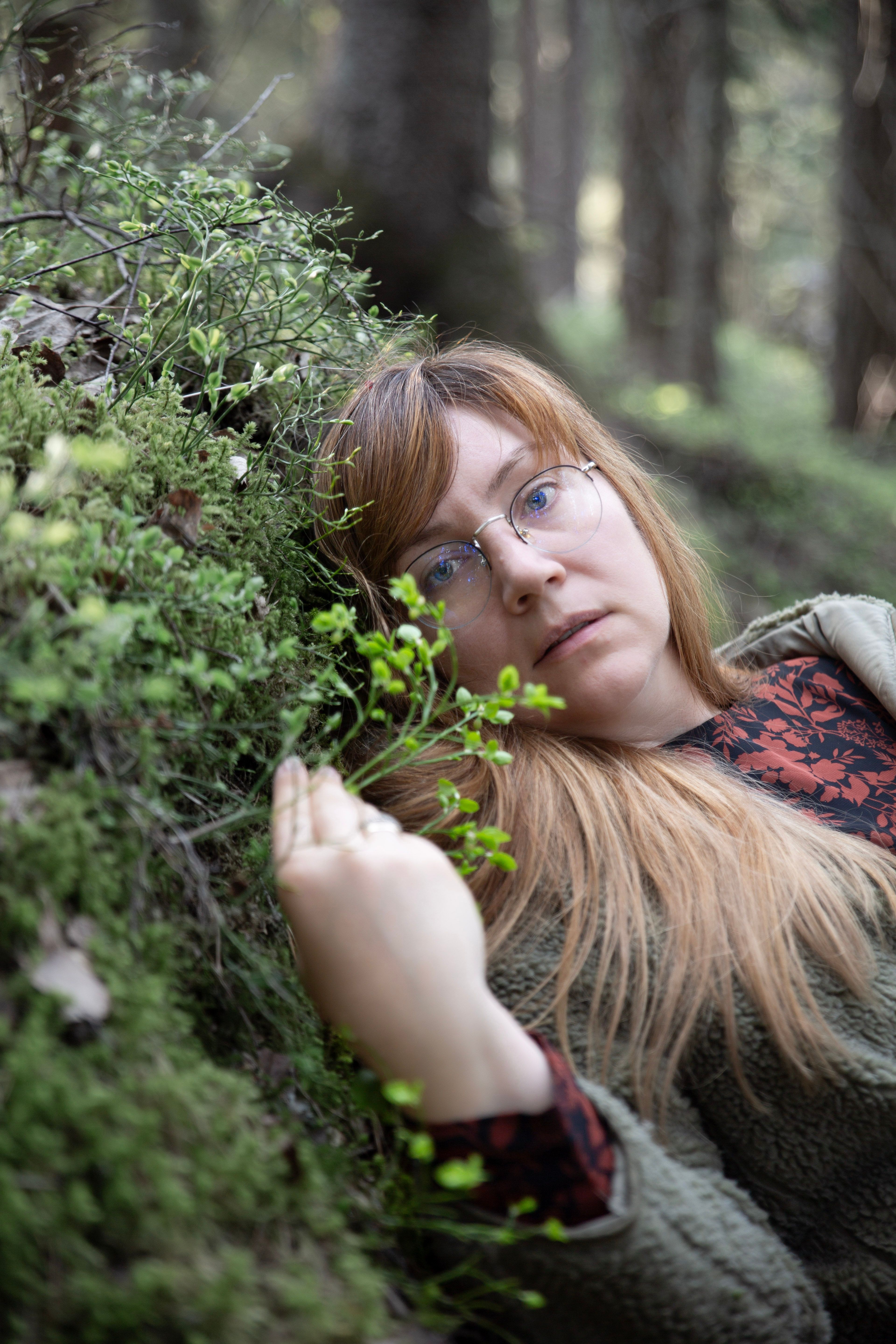 A woman with long blonde hair and eyeglasses lies peacefully in a lush forest setting.