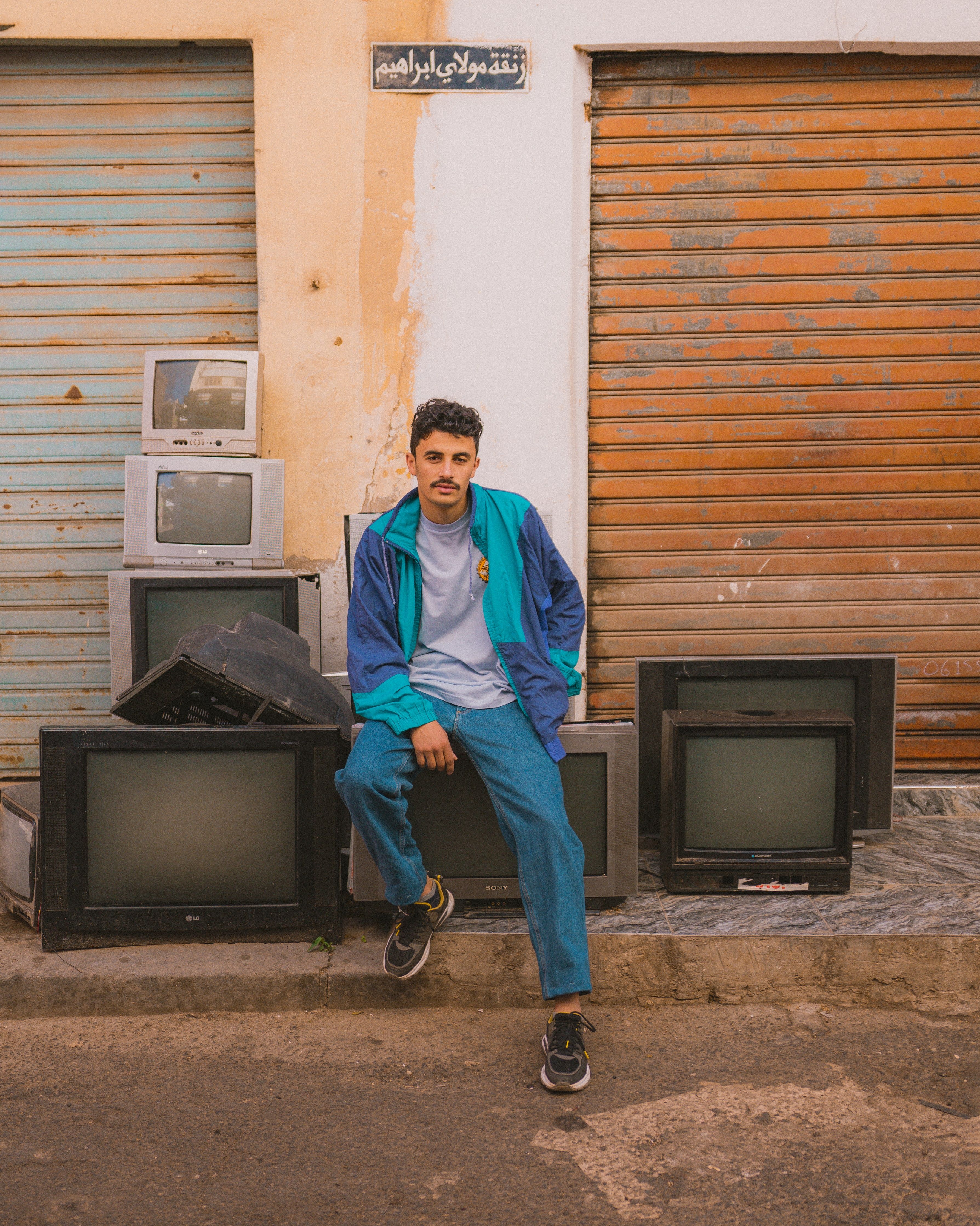 Young man in a vibrant jacket sitting by vintage TVs in an urban Moroccan street scene.