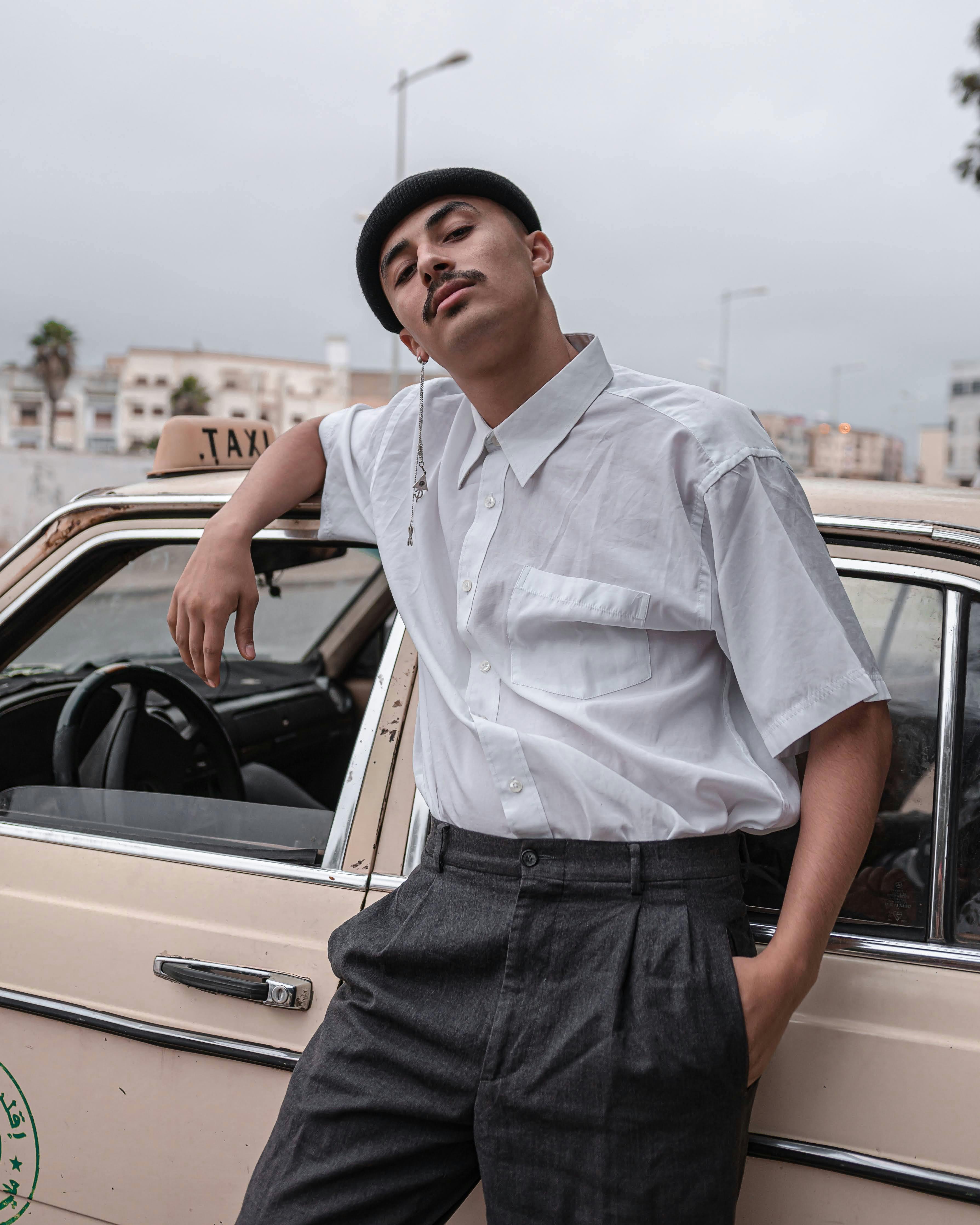 Young man in a white shirt and beret poses with confidence next to a vintage taxi.