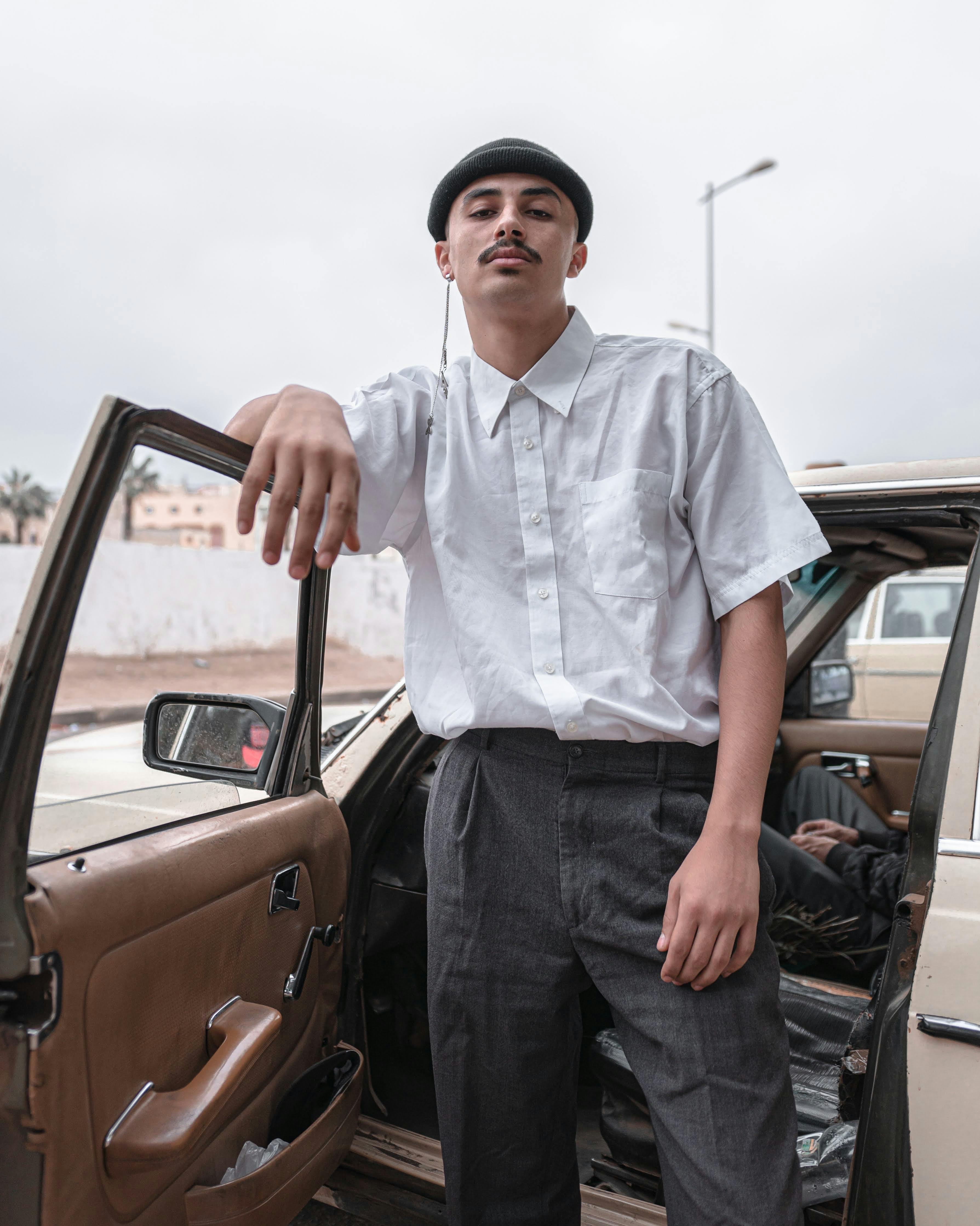 A stylish young man in a white shirt and beret poses by a classic car outdoors.