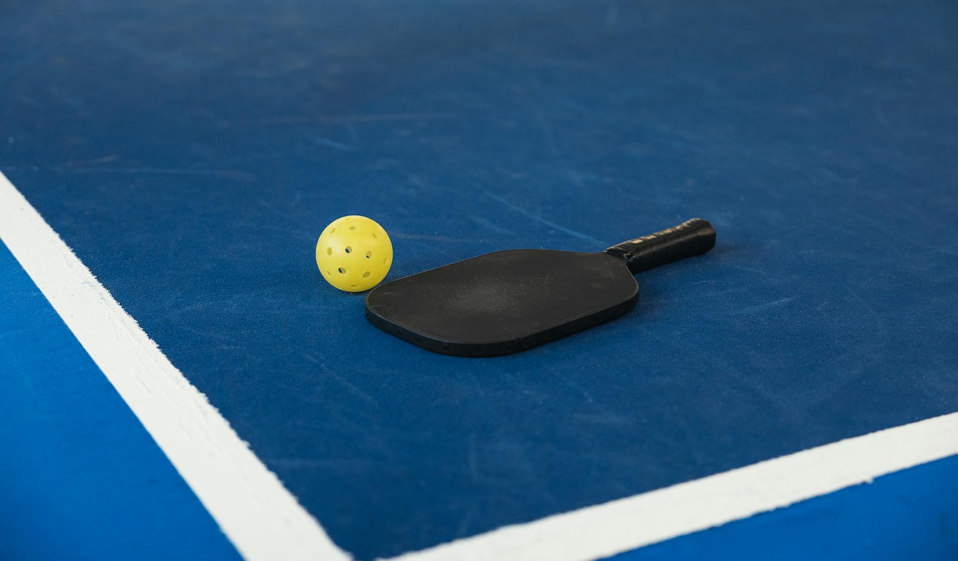 A pickleball paddle and yellow ball on a blue court corner, symbolizing sport and activity.