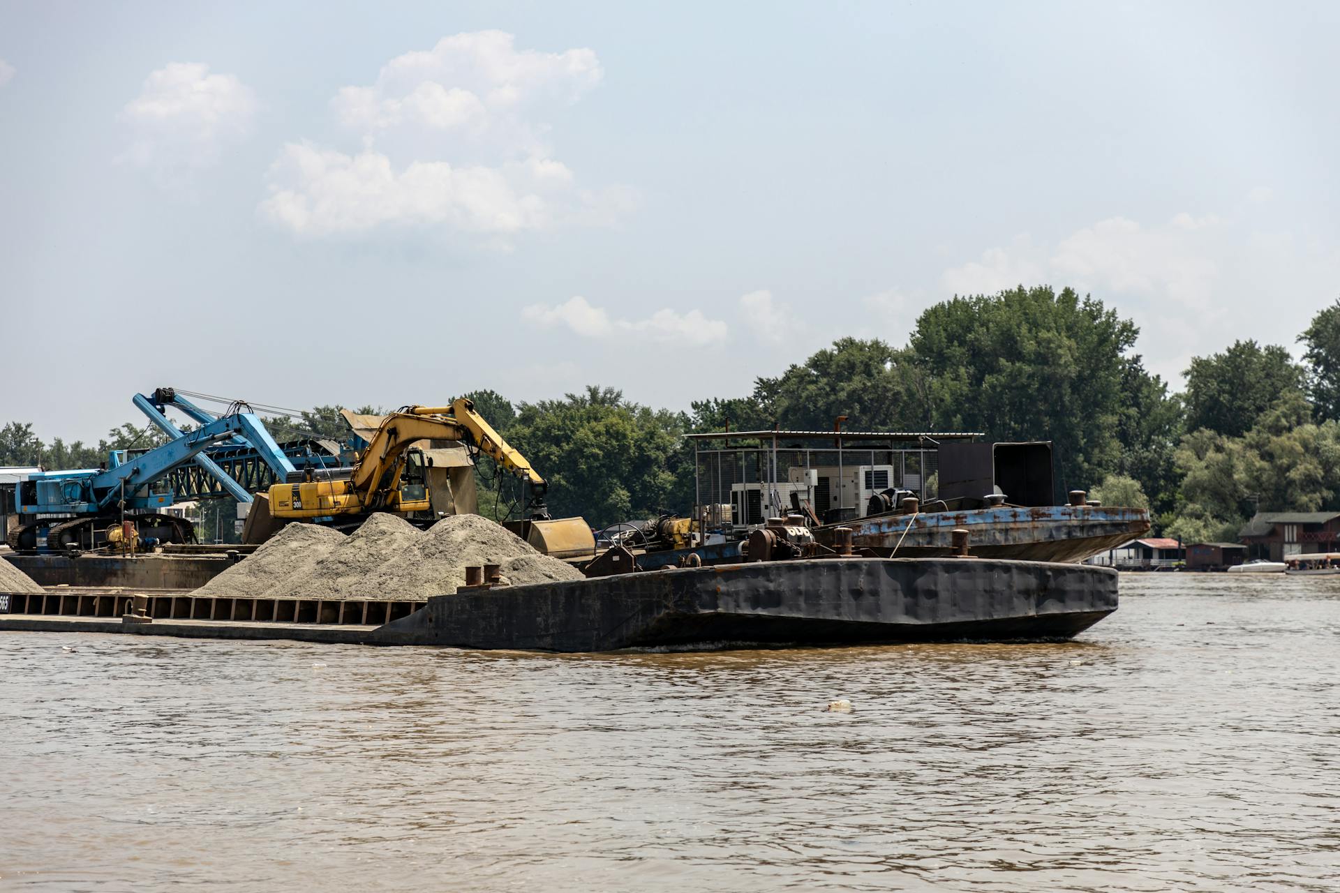 Barge with excavators and sand, working on a river under a bright sky.