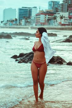 A young woman in a bikini enjoying a stroll on the beach with an urban backdrop.