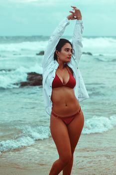 A woman in a red bikini with arms raised poses gracefully by the sea shore.