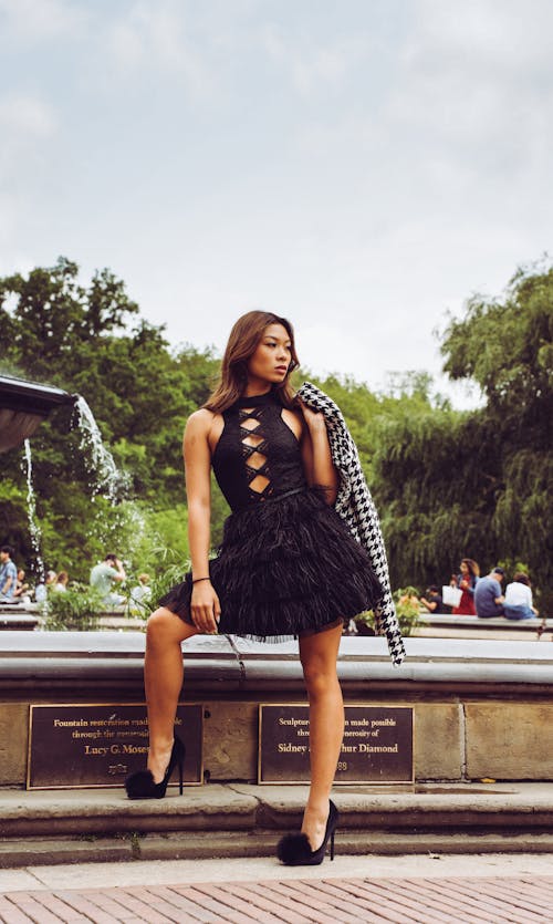 Photo of a Model Wearing a Black Dress Posing by a Fountain in a Park