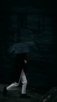 Blurred silhouette of a man with an umbrella running along a dark city street during rain.