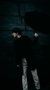 A man poses with an umbrella in a dark, moody setting, showcasing fashion and style.