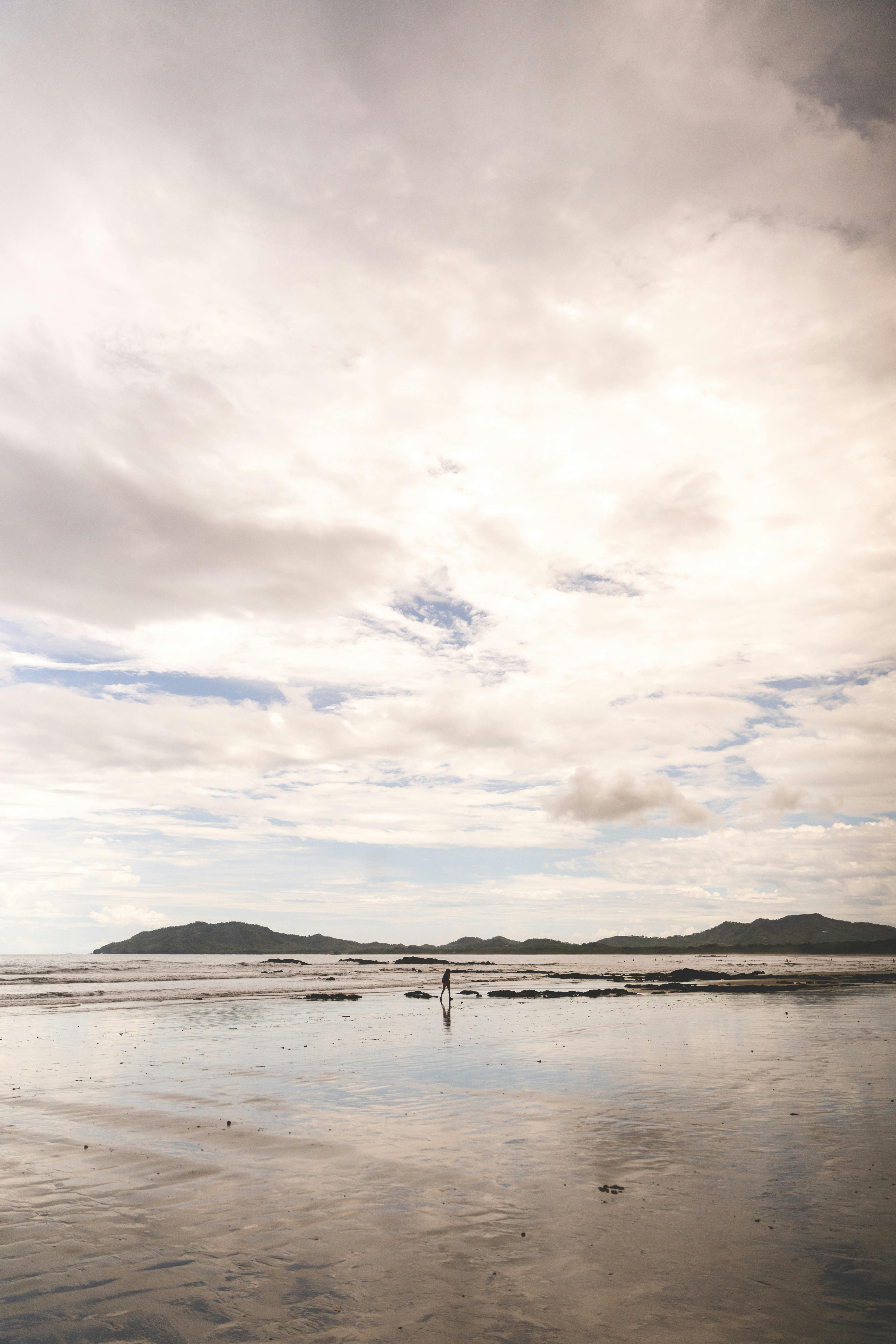 Serene beach scene featuring a cloudy sky and distant mountains reflecting in calm ocean waters.