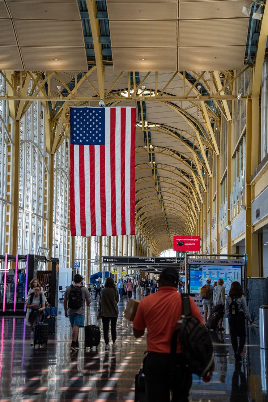 Travelers walk through a busy airport corridor beneath a large American flag