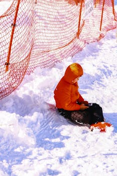 A child in an orange jacket sits in the snow holding a cellphone near a safety fence.