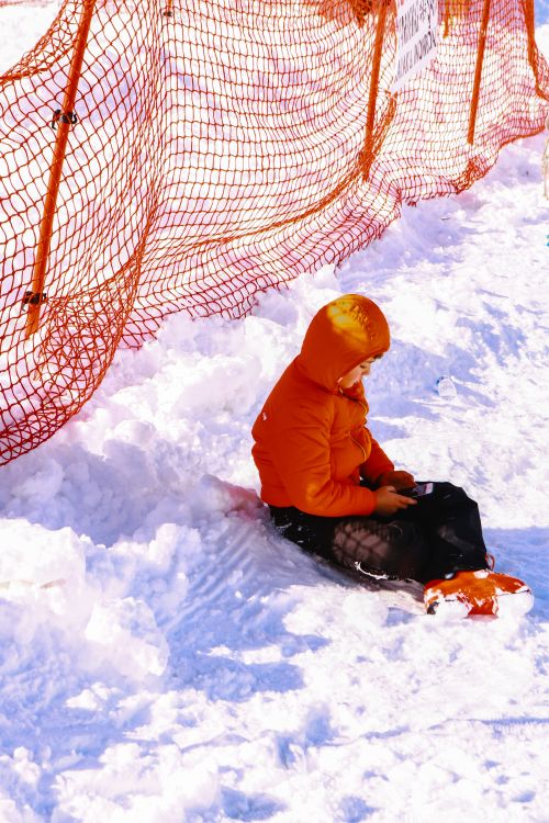 Free A child in an orange jacket sits in the snow holding a cellphone near a safety fence. Stock Photo