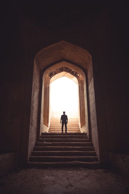 A solitary figure stands in a dramatic archway with steps leading to daylight.
