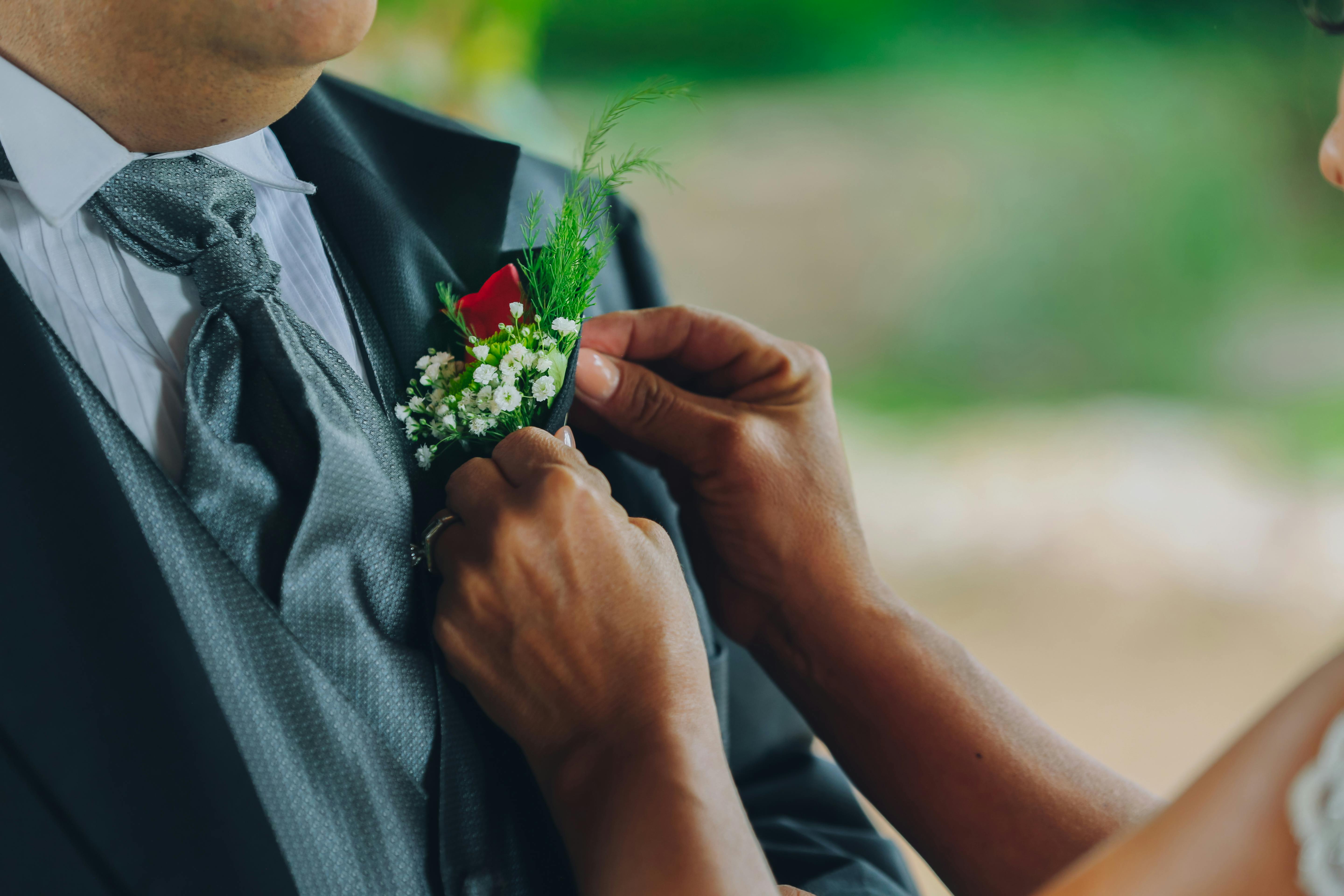 Close-up of a bride adjusting the groom's boutonniere during an outdoor wedding ceremony.