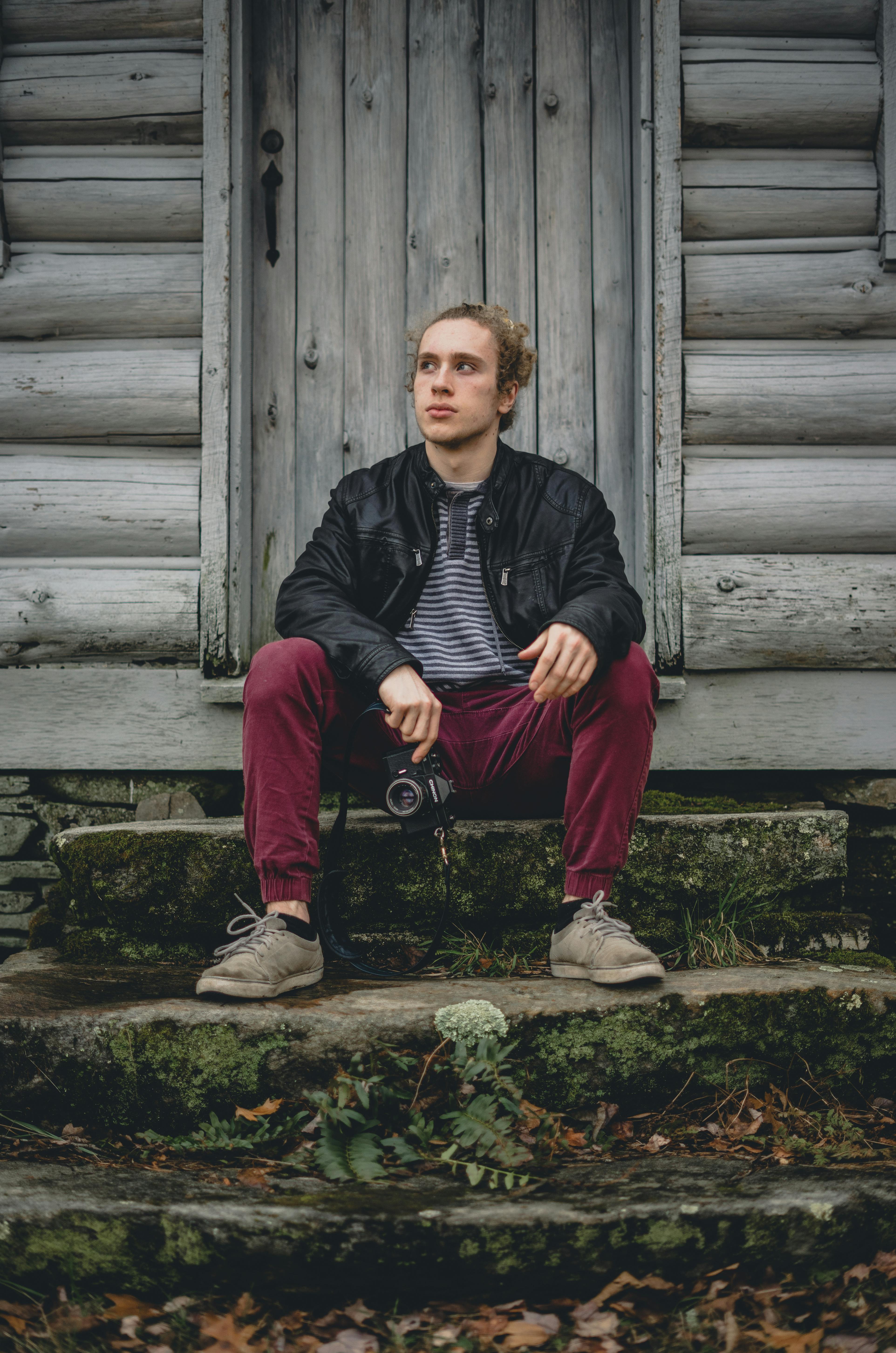 一名男子坐在小木屋的楼梯上，手持单反相机，神情轻松而沉思