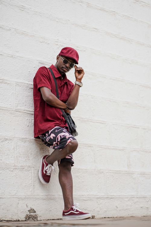Free Young man in sunglasses and red attire posing against a white brick wall with confidence. Stock Photo
