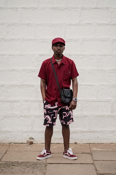 Stylish man in red shirt and hat standing against urban wall with crossbody bag.