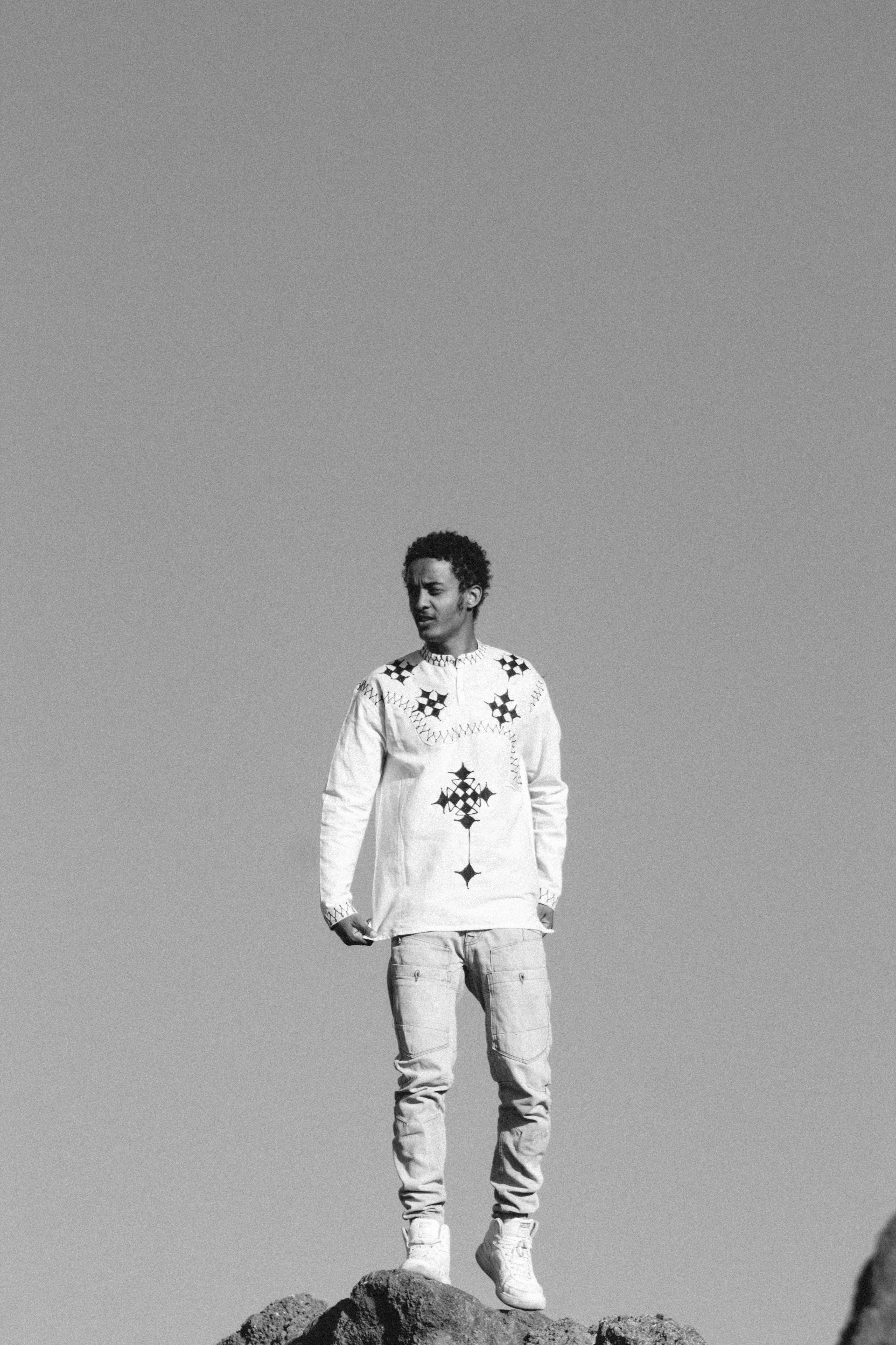 Black and white portrait of a man in traditional Ethiopian attire standing on rocks under a clear sky.