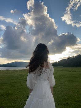 A woman in a white dress stands in a field, gazing at the dramatic clouds at sunset over the lake.