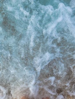 Top view of ocean waves crashing onto a sandy shore, creating white sea foam.