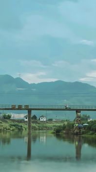 A peaceful countryside scene featuring a bridge over a serene river with distant mountains under an overcast sky.