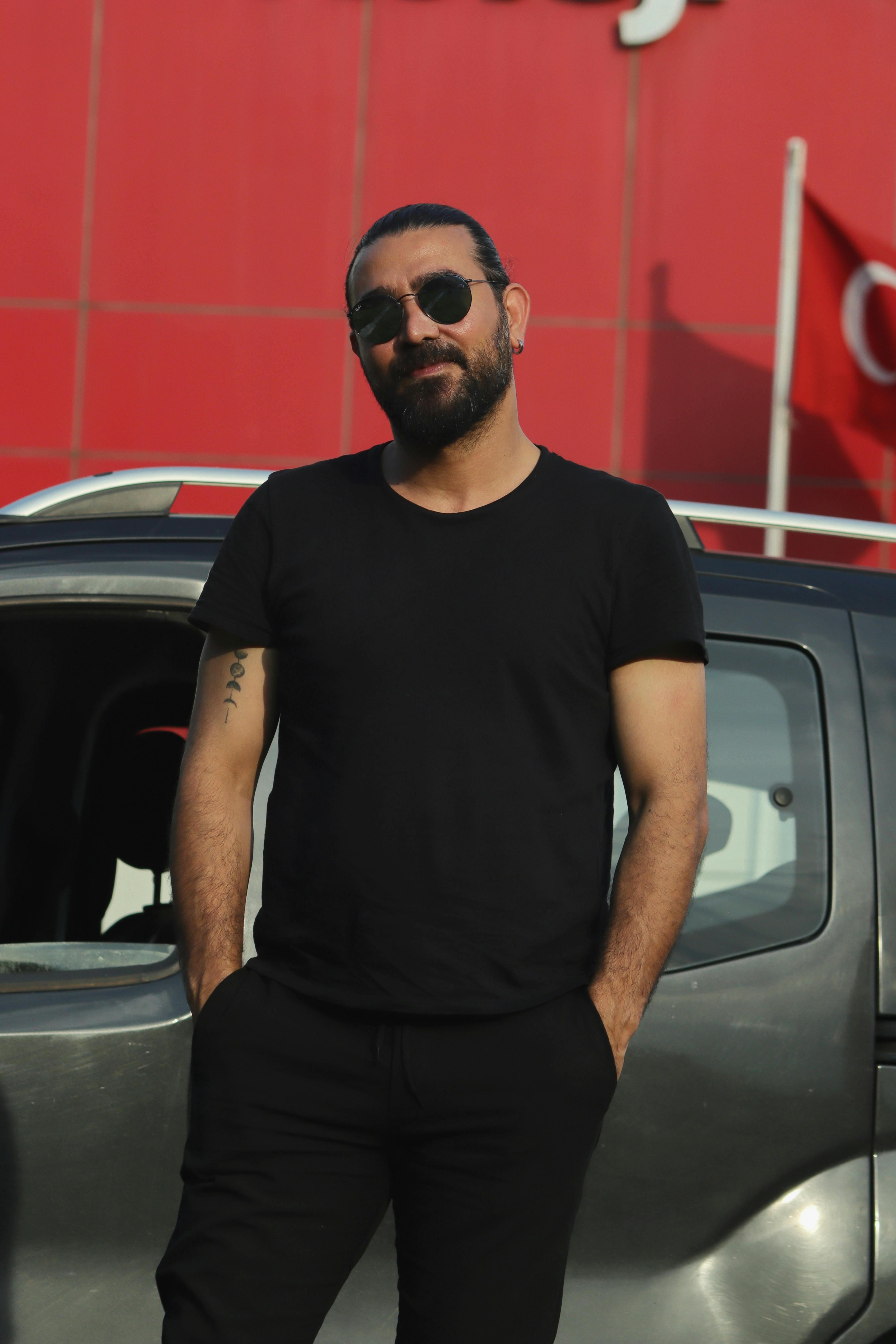 Man in black t-shirt and sunglasses stands by car with an urban backdrop.