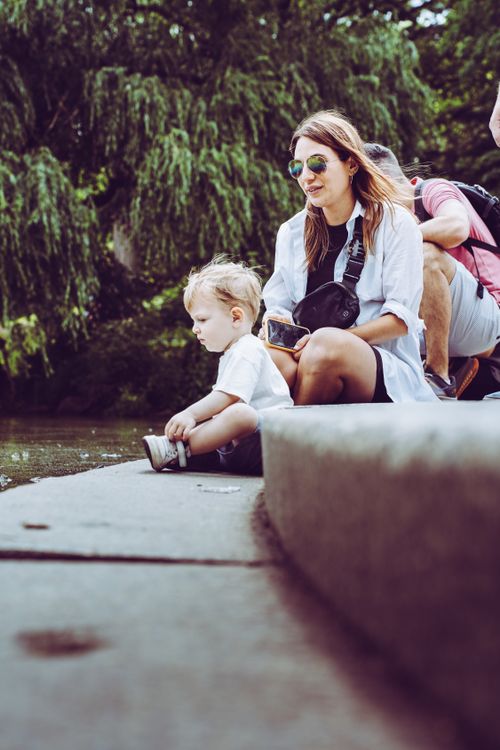 Free A woman and her child enjoy a peaceful moment sitting by a pond in a lush green park. Stock Photo