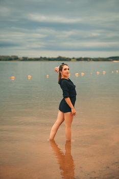 A woman in a black outfit standing in shallow water at a sandy beach, posing confidently on a cloudy summer day.