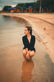 A woman in a black dress crouching by the lakeshore, creating a serene and moody atmosphere.