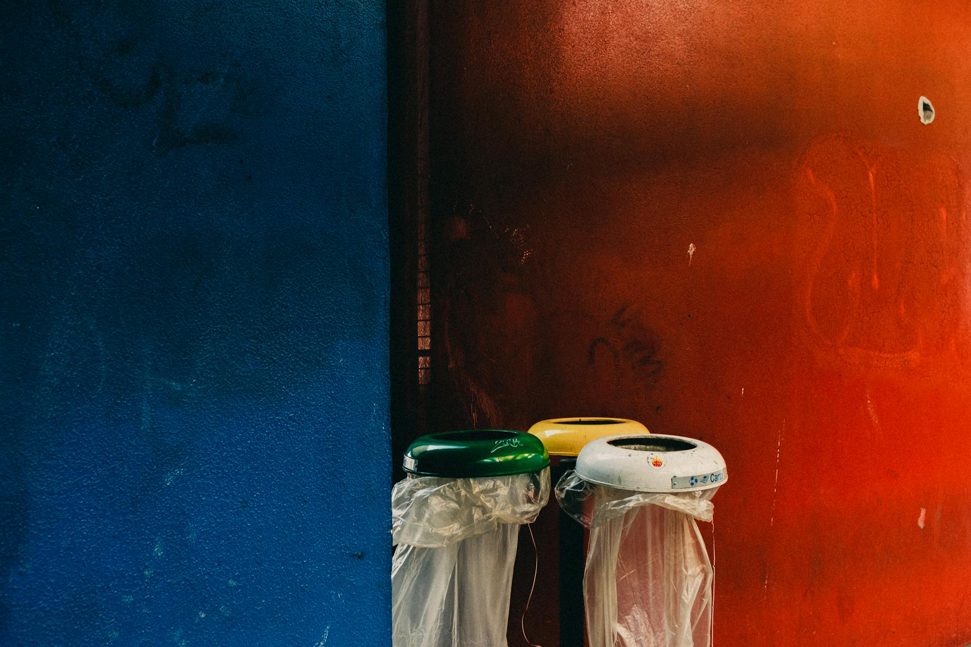 Plastic recycling bins against vibrant blue and red walls in an urban setting.