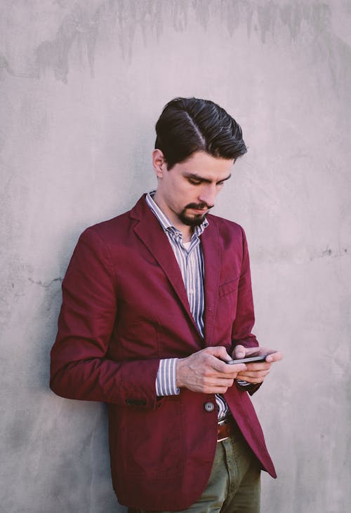 Free Man Wearing Maroon Blazer Leaning on Gray Concrete Wall While Using His Smartphone Stock Photo
