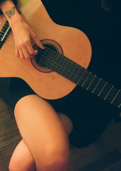 Close-up of a person playing an acoustic guitar, focusing on a tattooed hand and crossed legs.