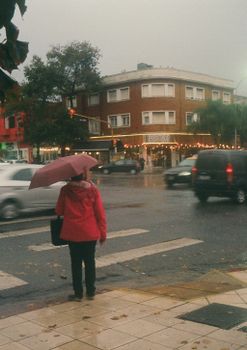 A woman with a pink umbrella stands at a rainy city intersection, surrounded by cars and buildings.
