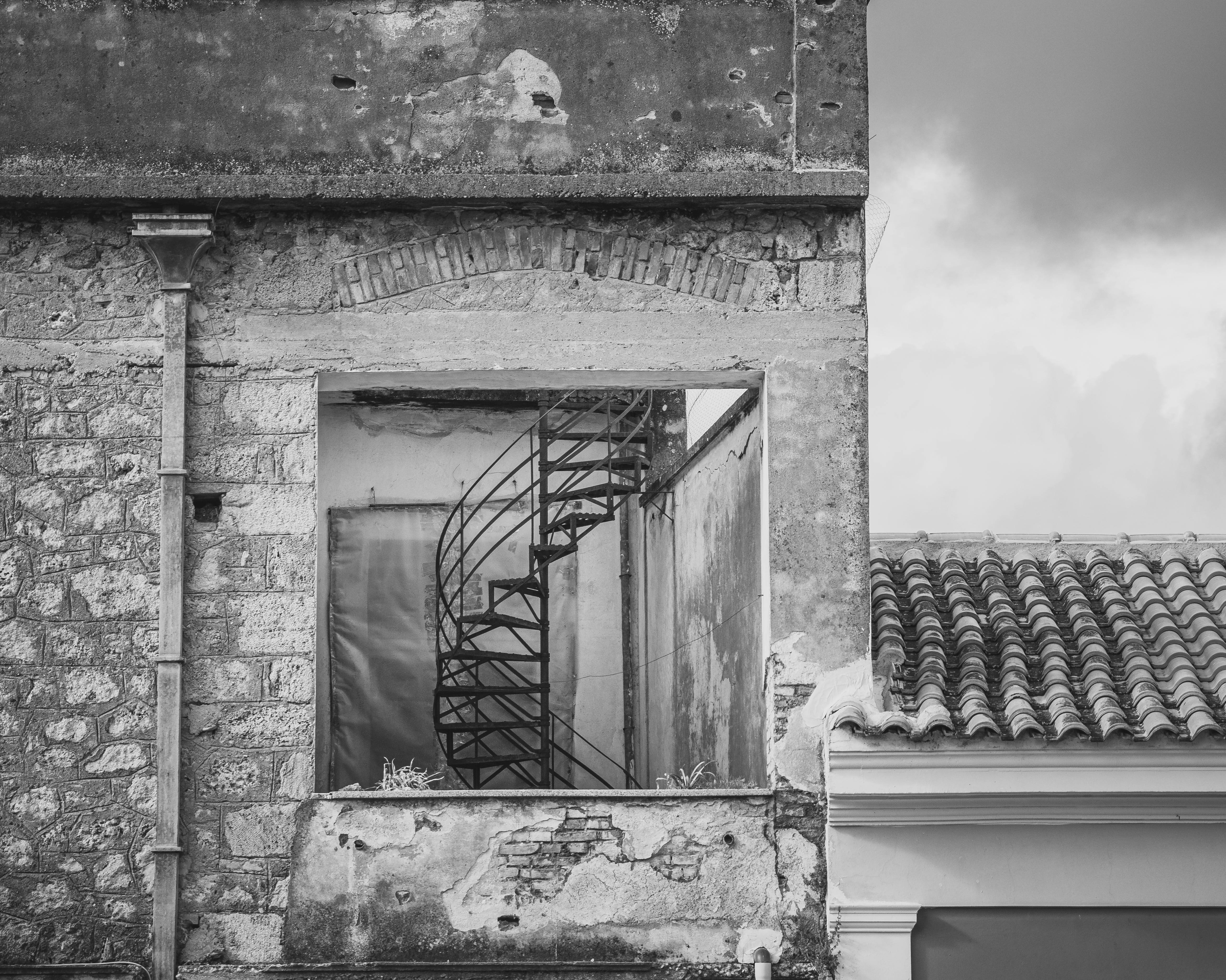 Black and white photo of an abandoned building with a spiral staircase visible through a window.