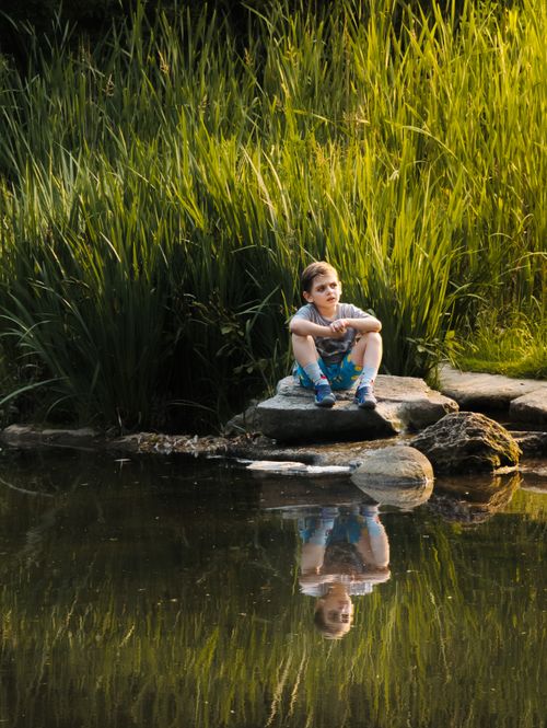 Free A teenager sits by a tranquil rural riverside surrounded by lush reeds, reflecting serenity. Stock Photo