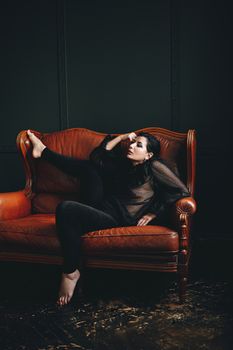 Fashionable woman in a black outfit poses elegantly on a vintage leather couch indoors.