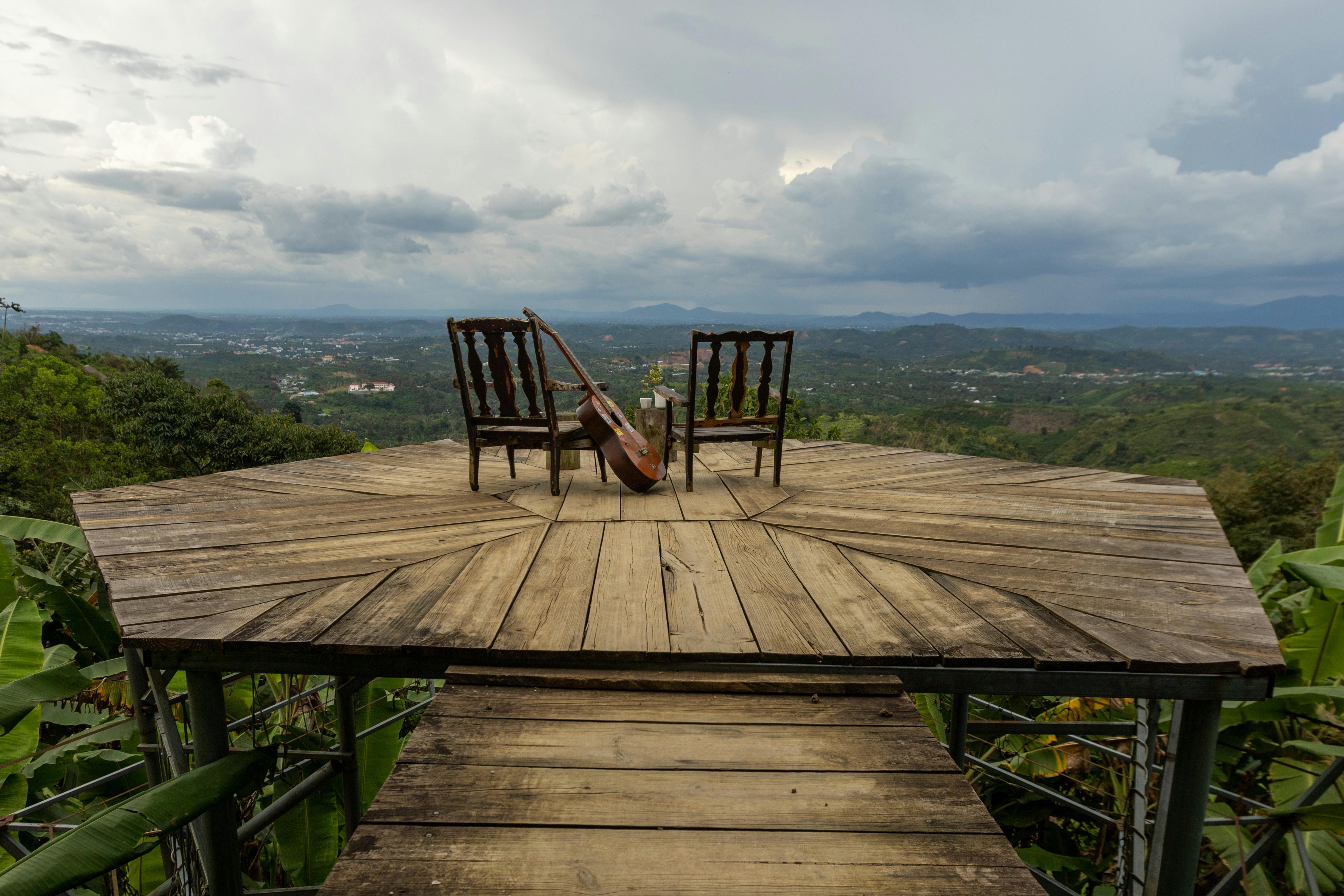 A wooden terrace with two chairs and a double bass overlooks a vast landscape under cloudy skies.