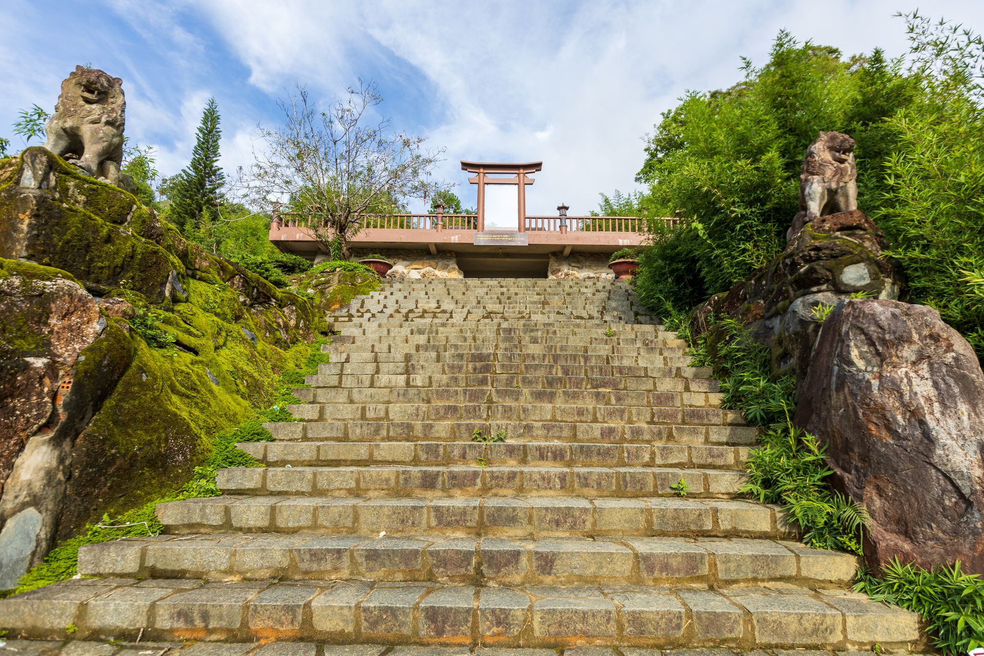 A serene pathway of stone steps flanked by lion statues leading to a scenic temple amid lush greenery.