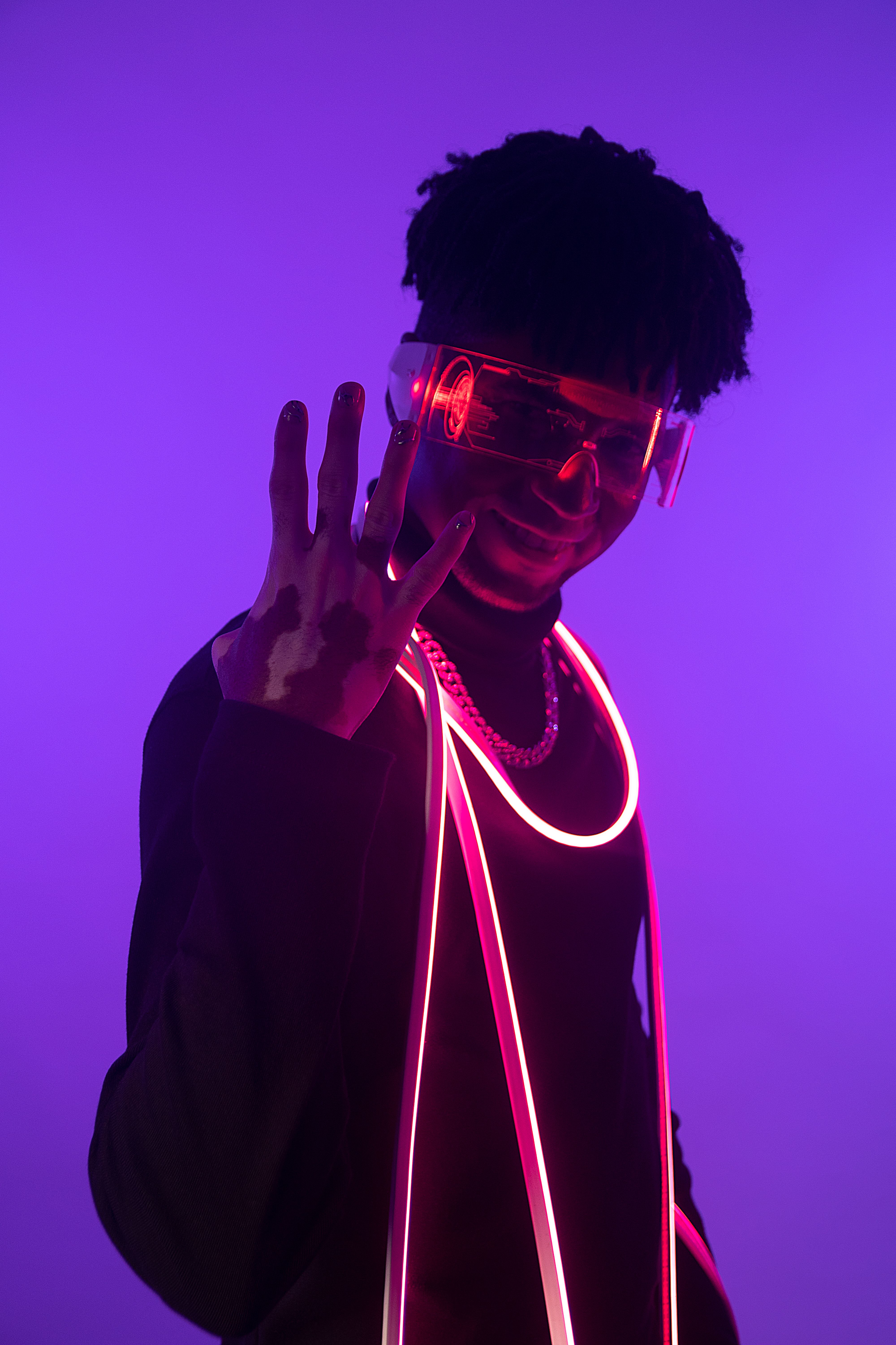 A fashionable adult man wearing eyeglasses and neon lights, standing against a vibrant purple background.