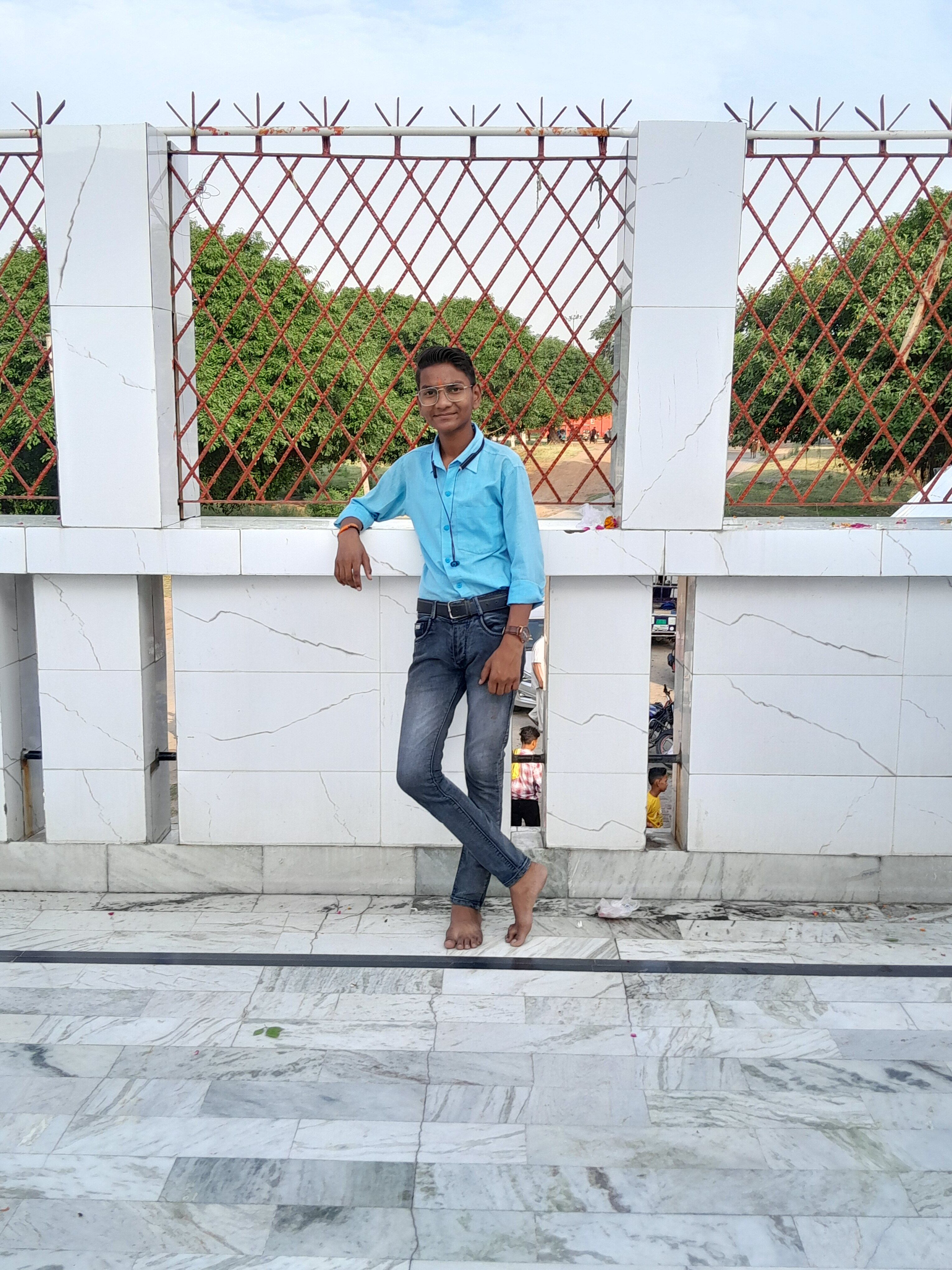Smiling young man in blue shirt leaning against a white wall with red latticework outdoors.