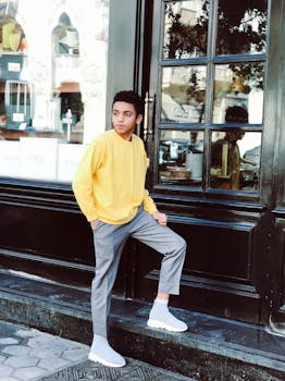 一位穿着黄色毛衣的时尚年轻人在城市街道的一栋建筑外摆姿势