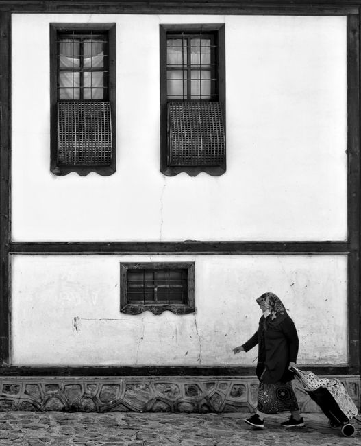 Black and white photo of an elderly woman walking by a historic building with wooden windows.